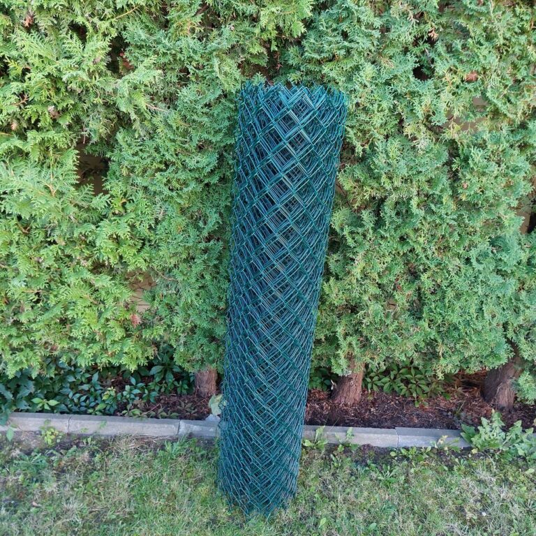 Zielona siatka ogrodzeniowa PCV zwinięta w rolkę, ustawiona na tle żywopłotu.